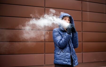 Vape teenager. Young pretty white caucasian girl in a blue jacket and hood smoking an electronic...