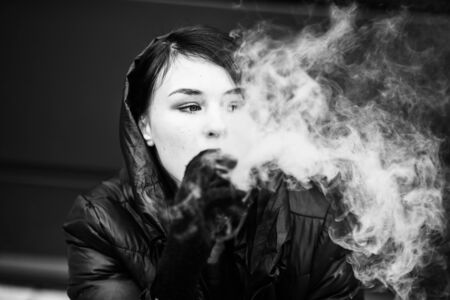 Vape teenager. Young pretty white girl smoking an electronic cigarette opposite modern background on the street in the winter. Deadly bad habit. Black and white.の写真素材
