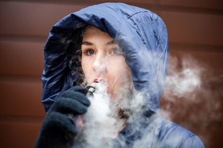 Vape teenager. Young pretty white caucasian girl in a blue jacket and hood smoking an electronic cigarette opposite modern brown background on the street in the winter. Deadly bad habit.の写真素材