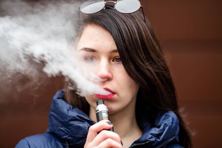 Vape teenager. Young pretty white caucasian girl in a blue jacket smoking an electronic cigarette...