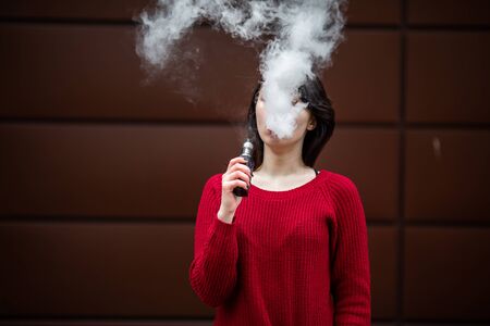 Vape teenager. Young pretty white caucasian girl in a red sweater smoking an electronic cigarette opposite modern brown background on the street in the winter. Deadly bad habit.の写真素材