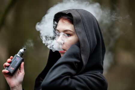 Vape teenager. Young pretty caucasian brunette girl smoking an electronic cigarette on the street in...