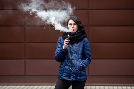 Vape teenager. Young pretty white caucasian girl in a blue jacket and glasses smoking an electronic cigarette opposite modern brown background on the street in the winter. Deadly bad habit.の写真素材