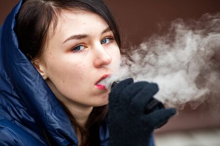 Vape teenager. Young pretty white caucasian girl in a blue jacket and hood smoking an electronic cigarette opposite modern brown background on the street in the winter. Deadly bad habit.の写真素材