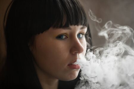 Vaping teenager. Young pretty white caucasian teenage brunette girl with blue eyes and problem skin smoking an electronic cigarette indoors. Deadly bad habit. Vape activity.の写真素材