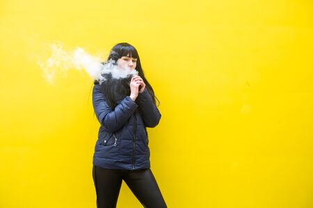 Vape teenager. Young pretty white caucasian brunette girl with blue eyes and in a blue jacket smoking an electronic cigarette opposite yellow wall on the street in the spring. Deadly bad habit.の写真素材