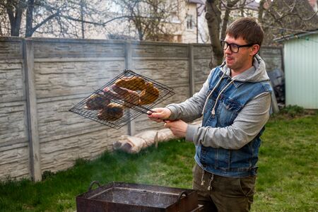 BBQ. Young bearded man in a denim jacket and in glasses cooks barbecue steak and sausages on the old grill in the backyard in the summer day.の写真素材