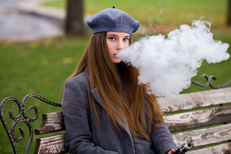Vaping teenager. Young pretty white caucasian teenage girl in gray beret smoking an electronic cigarette on the bench on the street in the autumn day. Bad habit. Vape activityの写真素材