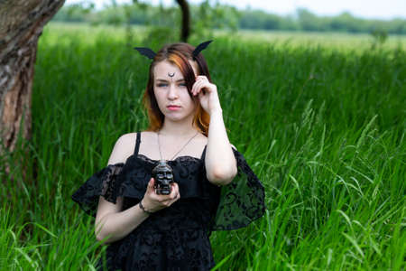 Young beautiful witch in black dress conducts a ceremony in a green forest on a summer day. Modern...