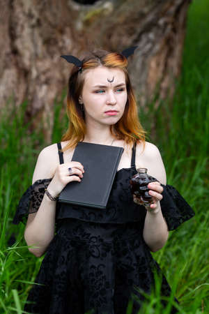 Young beautiful witch in black dress conducts a ceremony in a green forest on a summer day. Modern...