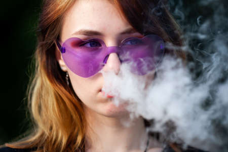 Vape teenager. Young pretty white caucasian girl in purple glasses on her head an electronic cigarette on the street in the summer. Deadly bad habit.の写真素材