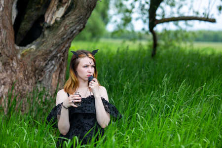 Young beautiful witch in black dress conducts a ceremony in a green forest on a summer day. Modern...