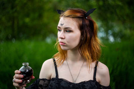 Young beautiful witch in black dress conducts a ceremony in a green forest on a summer day. Modern...