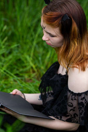 Young beautiful witch in black dress conducts a ceremony in a green forest on a summer day. Modern witchcraft.の写真素材