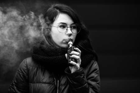 Vape teenager. Young pretty white caucasian girl in a jacket and glasses smoking an electronic cigarette opposite modern brown background on the street in the winter. Deadly bad habit. Black and whiteの写真素材