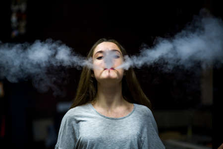Vaping teenager. Young pretty white caucasian teenage girl with problem skin smoking an electronic cigarette indoors. Deadly bad habit. Vape activity.の写真素材