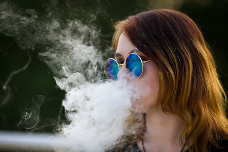 Vape teenager. Young pretty white caucasian girl in glasses on her head an electronic cigarette on the street in the summer. Deadly bad habit.の写真素材