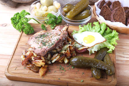 Fried pork with potatoes, egg and pickle cucumber on a wooden boardの写真素材