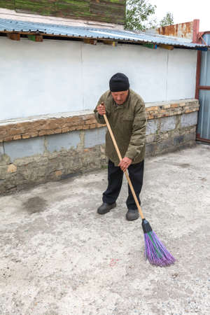 An elderly man sweeps the yard near his house. Household work in the village.の写真素材