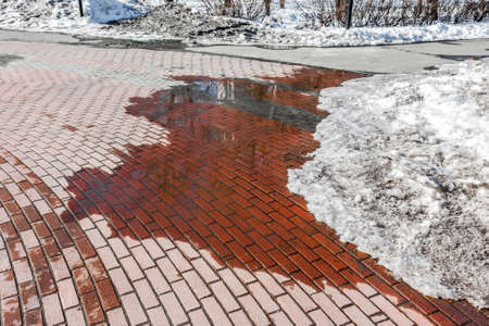 A puddle on the cobblestone sidewalk from melting snow. A sunny spring day.の写真素材
