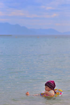 A little girl in a bandana floats on an inflatable ring alone in the sea against a background of blurry mountains and clouds. Side view, soft lilac background for copy space. Vietnam, Nha Trang.の写真素材