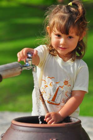 A little girl in a yellow T-shirt turned on a water tap in the park against a background of green grass. Water flows into an earthenware jug. Selective focus.の写真素材