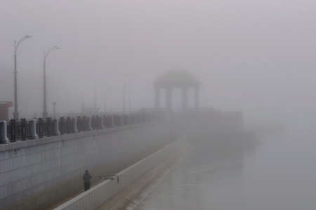 City embankment on a foggy morning during seasonal flooding. Lonely fisherman with a fishing rod. The silhouettes of the lanterns and the rotunda are heavily blurred in the fog.の写真素材