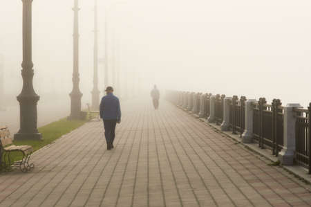 Thick fog on the Amur river embankment. Figures of people from the back. Street lights, benches and a fence. City of Blagoveshchensk, Russia.の写真素材