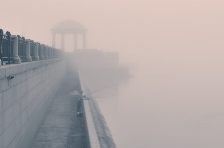 Silhouette of a fisherman on the embankment in the early morning fog. The outlines of the rotunda are blurred in the background. Before dawn.の写真素材