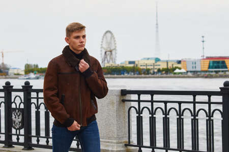 A European-looking guy in a warm brown jacket walks along the city embankment. Beyond the river, in blur, you can see the Ferris wheel, buildings and a TV tower on the Chinese coast. Selective focus.の写真素材