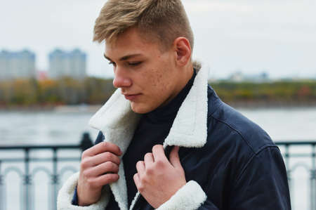 Portrait of a young Caucasian blond guy in a warm leather black jacket. The guy lifts the fur collar with both hands. Skin acne. In a blur in the background, the river, forest and buildings.の写真素材