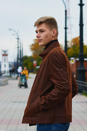 Portrait of a young man in a brown leather jacket on the autumn river embankment. In the background, a lighthouse and yellow foliage of trees are in blur. Skin acne. Selective focus.の写真素材