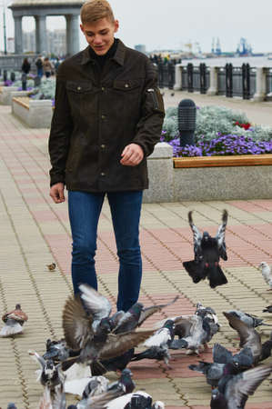 A guy in a warm jacket walks along the city embankment with pigeons against the backdrop of the rotunda. Blur the background. Selective focus. Blagoveshchensk, Russia.の写真素材