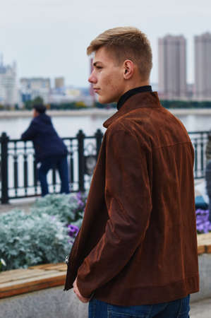 Fashion lifestyle. A young man in a brown leather jacket and jeans on the city embankment. The country is China across the river. Acne on the face. Blagoveshchensk, Russia.の写真素材
