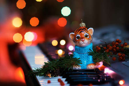 Christmas tree toy of a handsome tiger in a sweater on the piano keys. Colored lights of an electric garland. Branches of a Christmas tree and rowan berries. Happy New Year. Selective focus.の写真素材