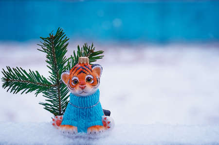 A tiger cub in a blue sweater with a branch of a Christmas tree sits in the snow outdoors....