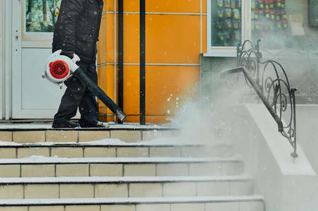 A janitor cleans snow from the stairs next to the shop with a wind blower. Lumps of snow and snowflakes in the air. Snow cloud. Work outdoors in winter. Snow removal after a snowfall. Selective focus.の写真素材