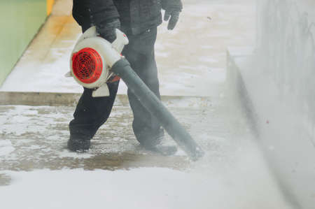 A Street cleaner removes snow from the sidewalk with a wind blower. Lumps of snow and snowflakes in the air. Snow cloud. Work outdoors in winter. Selective focus.の写真素材