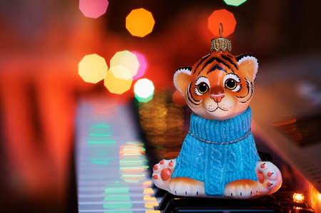 An orange tiger cub in a blue sweater sits on a musical synthesizer. In the background the lights of an electric Christmas garland. Reflection of light of lights. Greeting card. Selective focus.の写真素材