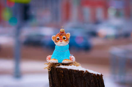 A toy tiger in a blue sweater is a symbol of the new 2022 year according to the Chinese calendar. Christmas tree toy on a city street on a tree stump. Traffic lights, cars and buildings in strong blurの写真素材
