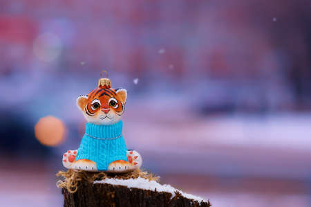 Tiger cub in a blue sweater on a stump in winter. Toy for the Christmas tree. Snowing. City street with transport in strong blur. Tiger is the symbol of 2022 on the Chinese calendar. Selective focus.の写真素材