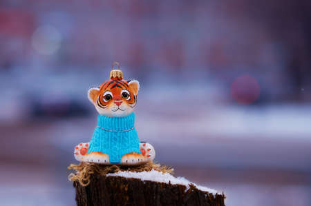 Tiger cub in a blue sweater on a stump in winter. Toy for the Christmas tree. Snowing. City street with transport in strong blur. Tiger is the symbol of 2022 on the Chinese calendar. Selective focus.の写真素材