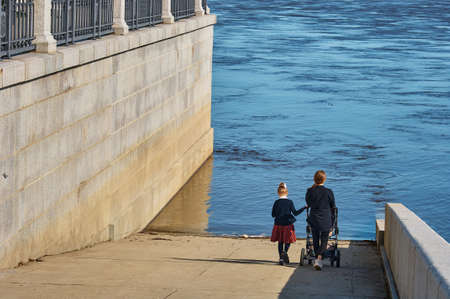 Two young girls with a baby in a stroller go down to the water along the descent of a granite embankment during a flood. Summer sunny day.の写真素材