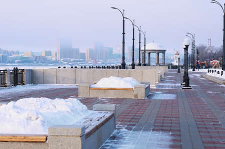 A bench with a flower bed in winter, filled with a snowdrift. Rotunda and lighthouse on the embankment. Blurred in the background is the city of Heihe, China. Blagoveshchensk, Russia.の写真素材