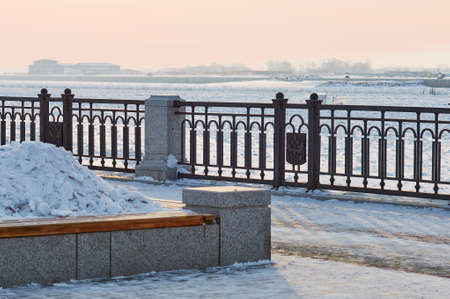 A bench with a flower bed in winter, filled with a snowdrift. Promenade of the city embankment. The concept of dual use. Blurred in the background is the city of Heihe, China. Blagoveshchensk, Russia.の写真素材