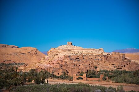 Ancient kasbah full of people lays between green oasis and hills with mountains background