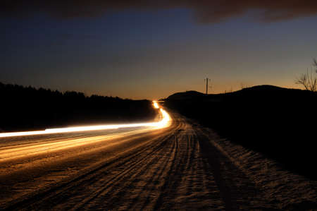 An asphalt road at dusk with the headlights of passing cars at a slow shutter speed. Night view with long passing headlights.の写真素材
