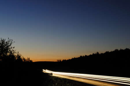 An asphalt road at dusk with the headlights of passing cars at a slow shutter speed. Night view with long passing headlights.の写真素材