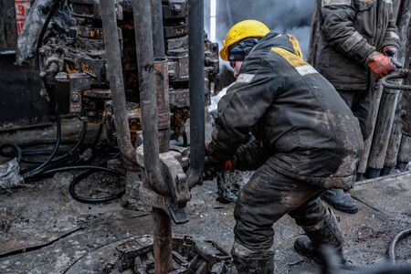 Two working drillers in a uniform, in a helmet and goggles, install drill pipes after lifting them...