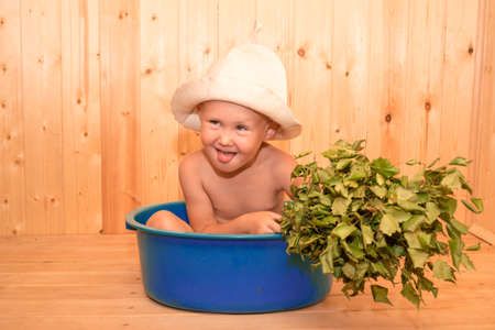 A child with a broom is sitting in a tub at the site. Traditional Russian bath. Funny hat, wellness treatments, little boy with bath hat and broom.の写真素材